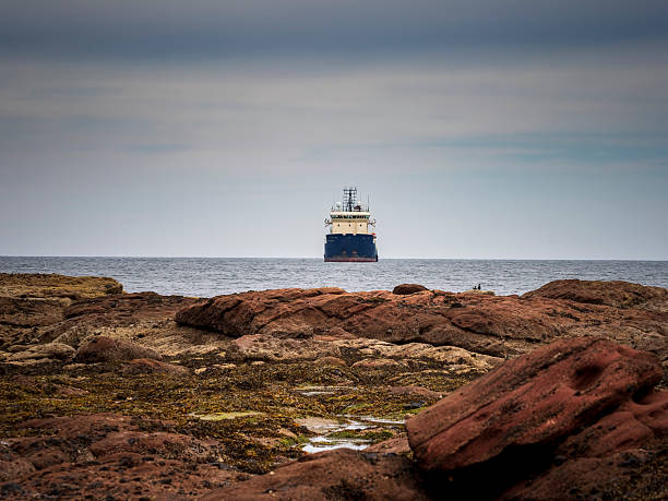 Blue ship sailing past a rugged rocky coastline under a cloudy sky