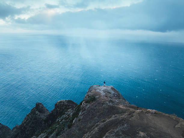 A lone hiker stands on a narrow cliff ridge beneath Miradouro da Garganta Funda on the island of Madeira, looking out over the vast Atlantic Ocean. Low clouds and soft light create a calm, moody coastal atmosphere.