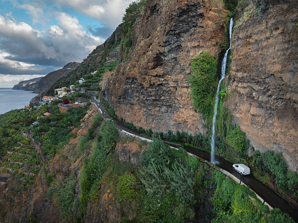 Aerial view of a car driving beneath Cascata dos Anjos on Madeira’s south coast, where a narrow cliffside road passes directly under a waterfall spilling from the rocks above.