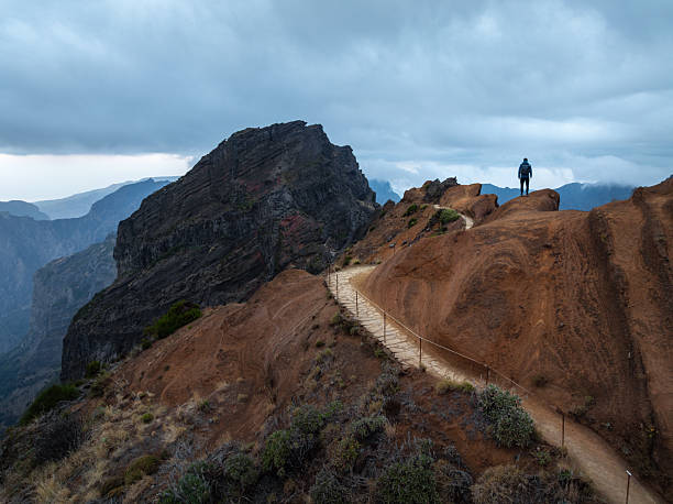A lone hiker stands on a narrow mountain ridge above steep volcanic cliffs, overlooking a vast and dramatic landscape (Pico do Arieiro, Madeira).