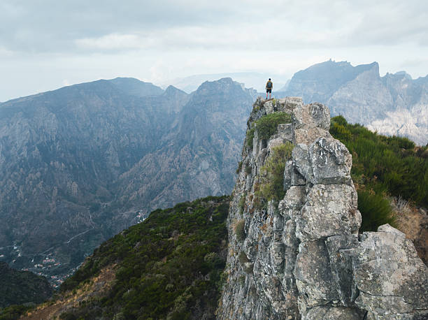 A lone hiker stands on a narrow rocky ridge high above deep valleys, looking toward Pico Grande in the central mountains of Madeira.