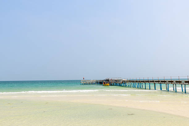 A long pier with blue supports extends from pale sand into clear turquoise water at Coconut Beach, Koh Rong, Cambodia. Gentle waves and a yellow boat create a tranquil, minimal seascape beneath a soft sky.