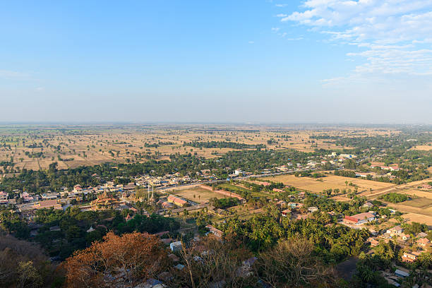 Wide view over golden agricultural fields, scattered trees, and rural settlements stretching across the plains near Phnom Sampov in Battambang province, Cambodia. The composition features patchwork farmland under a clear blue sky, evoking the dry season atmosphere of the Mekong region.