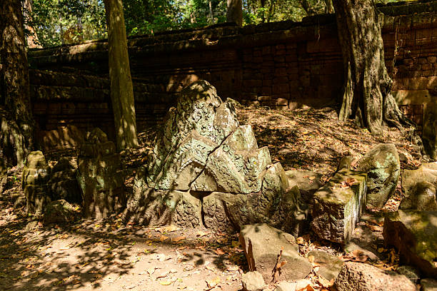 A moss-covered sandstone pediment with intricate carvings lies broken among fallen blocks on the forest floor near an ancient temple wall in Angkor, Cambodia. Dappled sunlight filters through the trees, highlighting the weathered textures and ornate Khmer details.