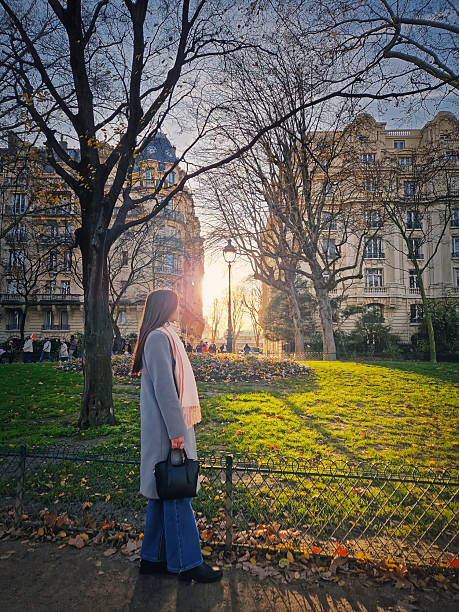 Beautiful young woman walking through a sunlit Parisian park, dressed in a grey coat while strolling past fallen autumn leaves towards the glowing sun through Haussmann style buildings