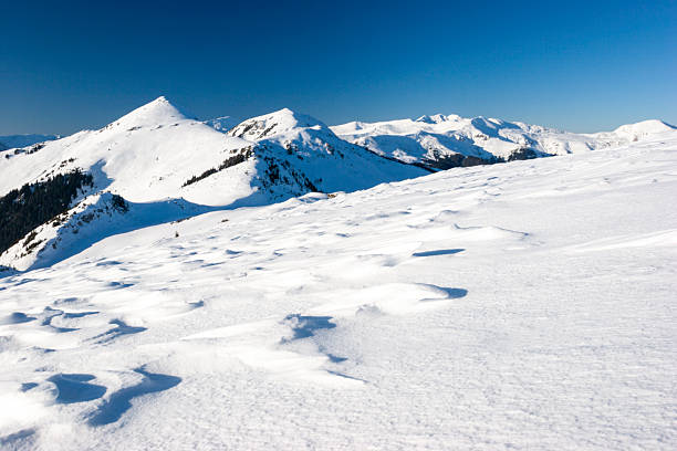 A wide-angle landscape shot of a massive, snow-blanketed mountain range beneath a brilliant, deep blue sky. The foreground highlights the unique wind-blown textures and ripples in the snow, leading the viewer's eye toward several sharp, sunlit peaks. The image evokes a sense of cold, quiet majesty and the vastness of the alpine wilderness, perfect for travel, adventure, and nature-themed projects.