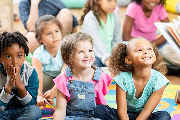 A daycare teacher sits with her students in front of her as she reads the group a story.  The children are each dressed casually in bright colours and focused on the story.
