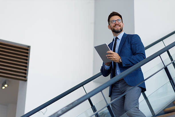istock Confident young businessman using digital tablet while walking upstairs in modern office building interior 2254793337