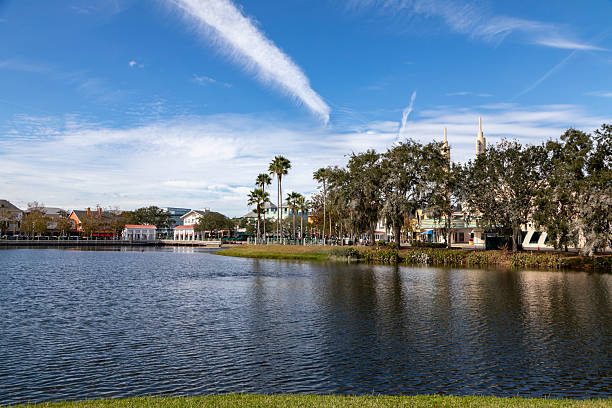 A wide scenic view of Celebration, Florida, showing the town center reflected in the calm waters of a central lake. Elegant buildings, palm trees, and distinctive architecture line the shoreline beneath a bright blue sky filled with wispy clouds.