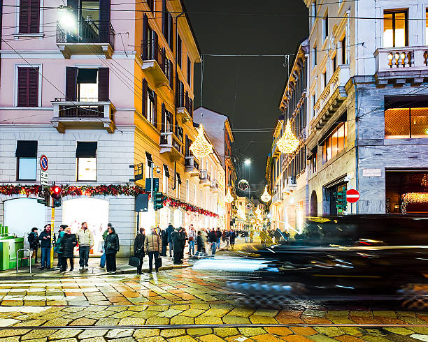 Night scene at Via Montenapoleone in Milan, showing a large crowd of people walking through the luxury fashion district. The street is decorated with elegant festive chandeliers and bright storefront lights, creating a vibrant urban atmosphere. This shot captures the motion of city life and high-end tourism in Italy's most famous shopping destination.