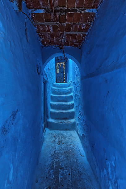 Narrow corridor beneath a red brick and wood beam framework supporting the houses above, ending at a dilapidated wooden door connecting the alleyways of the blue-painted medina. Chefchaouen-Morocco.