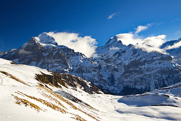 Snow-covered alpine mountain range under a deep blue sky with orographic fog. Rocky peaks in the Swiss Alps rise above rolling winter slopes covered in fresh snow, creating a dramatic high-altitude winter landscape. No people, suitable for travel, nature, and winter themes.