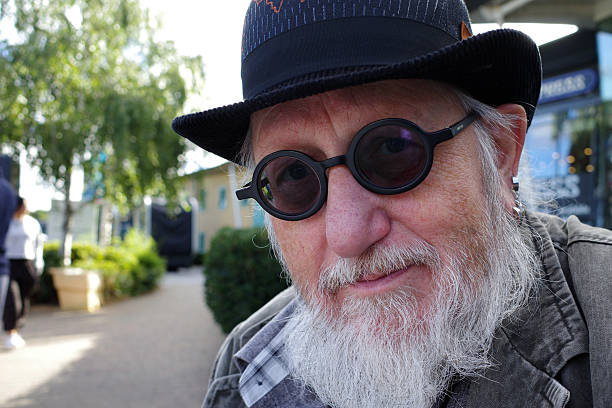 Senior man with a beard wears round black glasses and a bowler hat. He is looking directly at the camera with a slight smile. The background features a blurred outdoor area with trees and a patio
