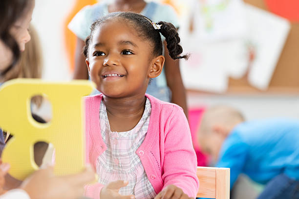 A happy young child is engaged in a classroom setting, learning with other children and holding a colorful letter.