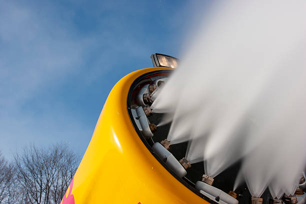 A dynamic, low-angle close-up of a bright yellow snow cannon blasting a powerful stream of artificial snow into a clear blue sky. The image captures the fine mist and heavy spray from multiple nozzles, illustrating the high-tech infrastructure used by ski resorts to maintain slopes. The vibrant yellow of the machine contrasts sharply against the sky, creating a clean, modern aesthetic suitable for travel brochures, winter sports articles, and industrial climate-control themes.