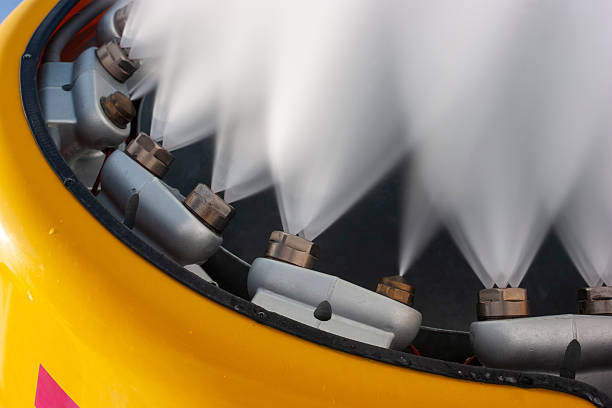 A dynamic, low-angle close-up of a bright yellow snow cannon blasting a powerful stream of artificial snow into a clear blue sky. The image captures the fine mist and heavy spray from multiple nozzles, illustrating the high-tech infrastructure used by ski resorts to maintain slopes. The vibrant yellow of the machine contrasts sharply against the sky, creating a clean, modern aesthetic suitable for travel brochures, winter sports articles, and industrial climate-control themes.