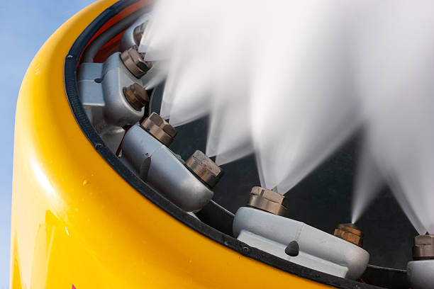 A dynamic, low-angle close-up of a bright yellow snow cannon blasting a powerful stream of artificial snow into a clear blue sky. The image captures the fine mist and heavy spray from multiple nozzles, illustrating the high-tech infrastructure used by ski resorts to maintain slopes. The vibrant yellow of the machine contrasts sharply against the sky, creating a clean, modern aesthetic suitable for travel brochures, winter sports articles, and industrial climate-control themes.