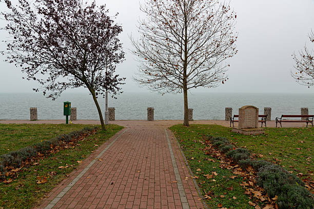 A walkway leads to the edge of Balaton lake. Two trees stand on either side. Benches are placed along the path. The sky is overcast and the water is calm.
