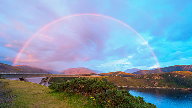 Kylesku Bridge spanning over water under a rainbow and colorful sky at sunset