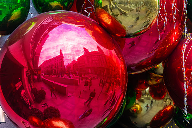 A cluster of oversized Christmas baubles—shining in gold, red, and green—adorns a festive corner of central Madrid. Their mirrored surfaces vividly reflect the surrounding architecture of Calle de Alcalá and the recently restored Centro Canalejas complex, capturing the essence of the city's holiday charm.