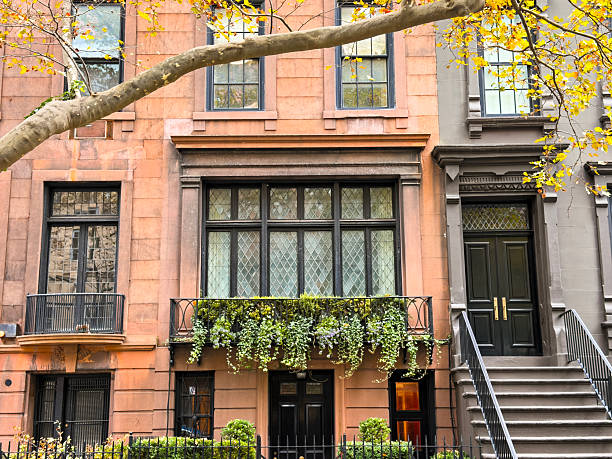 Facade and entrance of a classic Brooklyn brownstone townhouse in New York City, featuring traditional stoop stairs, black front door, large windows with wrought iron railings and flower boxes, framed by autumn tree branches and sidewalk greenery in warm daylight.Suitable for use in real estate and neighborhood guides, New York City lifestyle blogs, architecture and design features, urban living concepts and marketing materials highlighting historic Brooklyn residential streets.