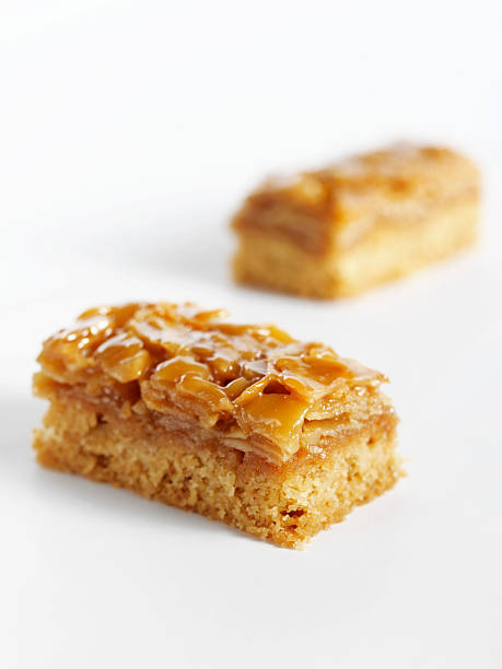 Vertical close up image of small rectangular caramel cookies topped with chopped nuts, arranged on a smooth white background. The minimal studio composition and soft lighting emphasize the golden color, texture, and glossy caramel surface, creating an appetizing dessert scene with ample copy space for text. This isolated sweet snack image is ideal for illustrating confectionery products, bakery items, and dessert concepts in both editorial and commercial layouts.