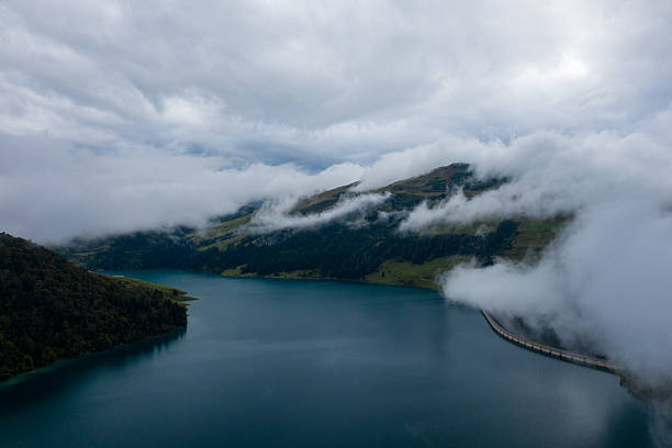 Clouds and mist drift across the calm blue waters of Roselend lake, with the curved concrete dam and forested alpine slopes partially obscured by low fog. The scene captures soft light, subtle textures, and the tranquil atmosphere of the French Alps.