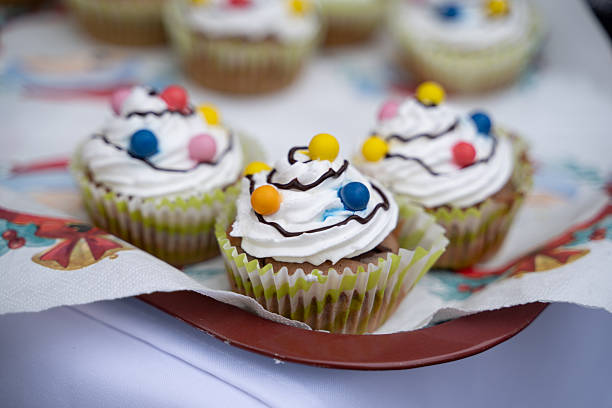 Cupcakes topped with swirls of whipped cream, drizzled chocolate, and vibrant candy balls are arranged on a festive tray with decorative napkins. The playful colors and textures create a joyful, celebratory atmosphere, perfect for parties, fairs, or holiday events.