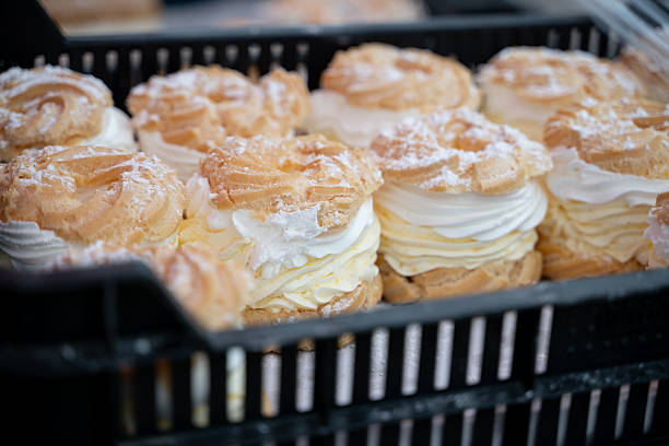 Rows of cream-filled choux pastries are neatly arranged in a black plastic tray, topped with powdered sugar. The delicate texture of the golden pastry contrasts with the rich, whipped filling. This sweet treat evokes a sense of indulgence, freshness, and traditional baking at a local market or bakery.