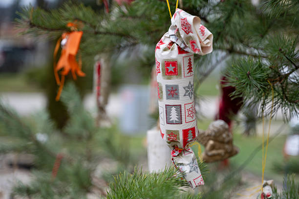 A handmade Christmas ornament wrapped in patterned holiday fabric hangs from a pine tree branch. The fabric features red and gray festive symbols like trees and stars, adding a rustic charm to the natural outdoor setting. The decoration reflects a cozy, DIY approach to holiday traditions.