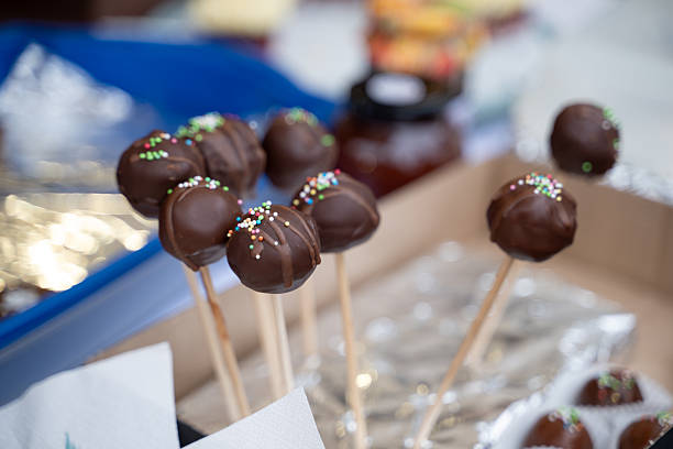 Chocolate-covered cake pops decorated with colorful sprinkles are arranged on wooden sticks, displayed in a cardboard box. The glossy coating and playful toppings make them visually appealing and festive. These sweet treats are perfect for parties, markets, or holiday celebrations.