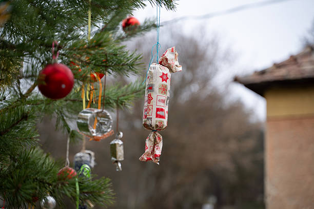 A handmade Christmas ornament wrapped in patterned holiday fabric hangs from a pine tree branch. The fabric features red and gray festive symbols like trees and stars, adding a rustic charm to the natural outdoor setting. The decoration reflects a cozy, DIY approach to holiday traditions.