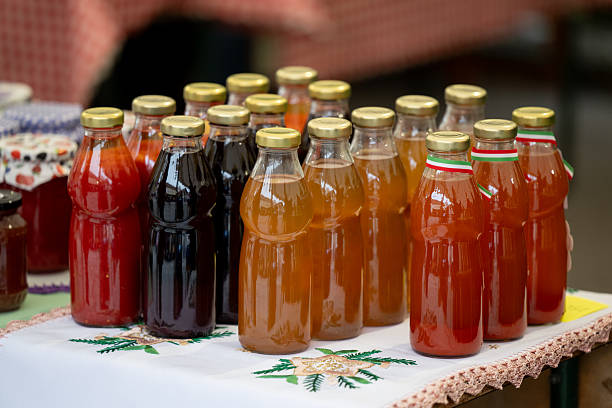 A variety of homemade fruit syrups in glass bottles with golden caps are arranged neatly on a table covered with an embroidered cloth. The vibrant colors range from deep red to amber and orange, indicating different flavors. The scene evokes a traditional market atmosphere and artisanal food culture.