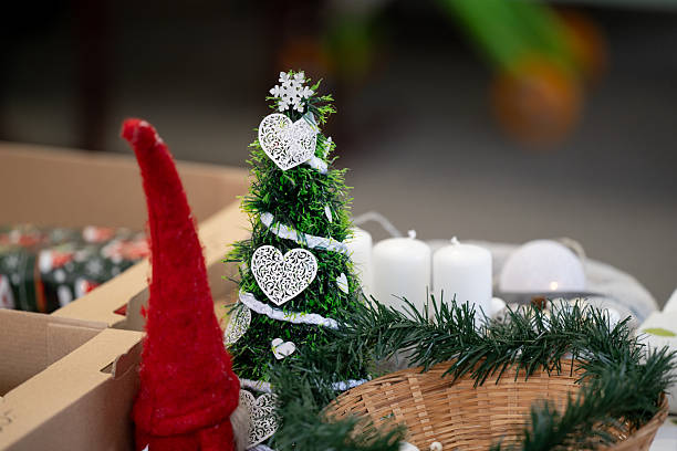 A small artificial Christmas tree adorned with white heart ornaments and ribbon stands beside white candles, a wicker basket, and a red felt gnome. The handcrafted decorations rest on a festive table, creating a cozy, artisanal holiday atmosphere. The scene reflects warmth, creativity, and seasonal cheer.