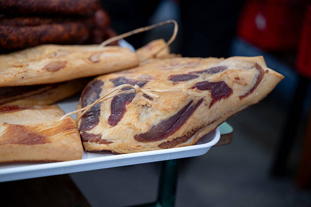 Thick slabs of cured pork belly with visible marbling and natural fat layers are displayed on a white tray at an outdoor market. Twine is tied through the meat for hanging or storage. The image showcases traditional preservation techniques and highlights the artisanal quality of local meat products.