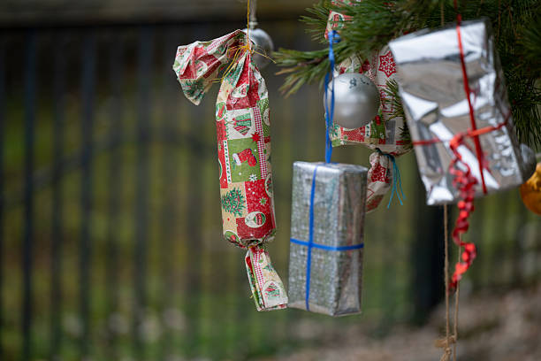 A handmade Christmas ornament wrapped in patterned holiday fabric hangs from a pine tree branch. The fabric features red and gray festive symbols like trees and stars, adding a rustic charm to the natural outdoor setting. The decoration reflects a cozy, DIY approach to holiday traditions.