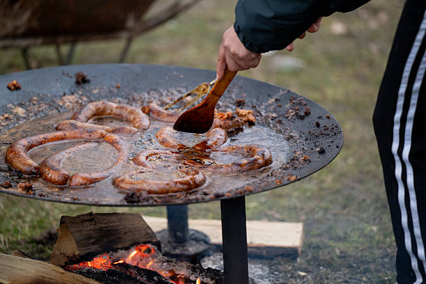 Coiled sausages are being grilled on a suspended metal pan above an open fire, with a person using metal tongs to turn the food. The outdoor setup and sizzling surface suggest rustic cooking or a communal barbecue. The scene captures the essence of traditional, fire-cooked meals in nature.