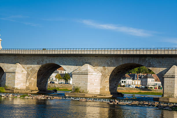 Historic stone bridge with wide arches spans the Loire river, reflecting in calm water under a clear blue sky. Traditional houses and greenery line the riverbank, highlighting the architectural heritage of La Charite sur Loire.