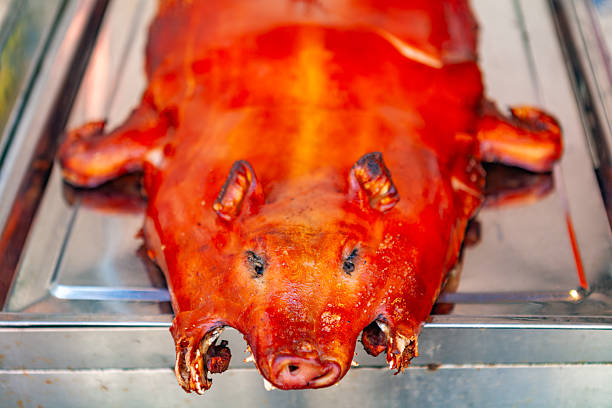 This close-up image showcases a whole roasted pig, likely a suckling pig, presented on a reflective silver or stainless steel tray. The pig's skin is a vibrant, glistening reddish-orange, suggesting a perfectly crispy texture from thorough roasting. Its head is prominent in the foreground, with visible snout, dark eye sockets, and slightly charred ears. The lighting highlights the rich, appealing color and texture of the cooked meat. The overall presentation is direct and inviting, evoking traditional festive cuisine or a display from a market or restaurant specializing in roasted meats.