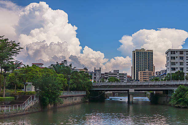 This image captures a vibrant cityscape where an urban river flows through the foreground, reflecting the light from above. A modern concrete bridge spans the river, connecting the lush green banks that are dotted with various buildings. On the left, a landscaped area features trees, steps with railings, and smaller structures. The right bank showcases a mix of architectural styles, including a prominent multi-story building and several other residential or commercial structures nestled among greenery. Above the urban landscape, a brilliant blue sky is adorned with large, fluffy white cumulus clouds that add a dramatic and expansive feel to the scene. The lighting suggests a bright, clear day, casting shadows and highlighting the textures of the water, foliage, and buildings.