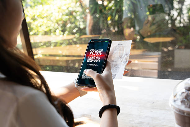 Woman holding smartphone scanning QR code with receipt in hand at cafe table near window. Concept of cashless payment, mobile banking, digital wallet, fintech, contactless checkout and spending.