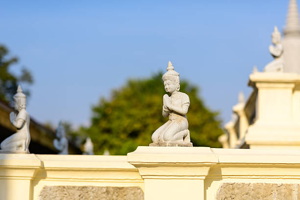 Kneeling guardian statue with traditional Khmer headdress and prayer gesture sits atop a pale balustrade in the Royal Palace grounds, Phnom Penh, Cambodia. Warm afternoon light enhances the sculpted details against a backdrop of greenery and blue sky.