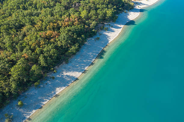 Aerial view of a tranquil pebble shoreline meeting clear turquoise water, bordered by dense green forest at Lac de Sainte-Croix on a sunny day.