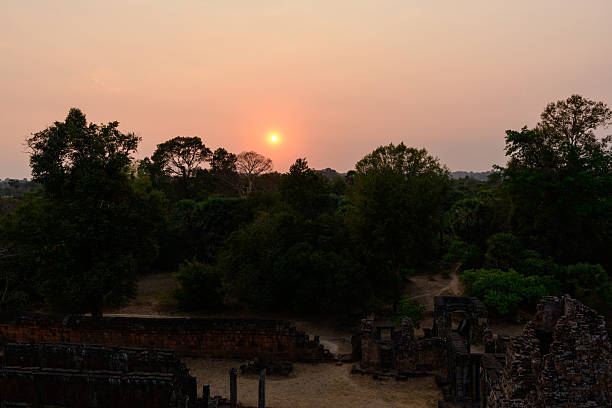 The setting sun casts a warm orange glow above dense tropical trees and the weathered stone ruins of Pre Rup temple in Cambodia. The tranquil scene features silhouettes of historic walls against a hazy sky, evoking the serene atmosphere of the Angkor region.