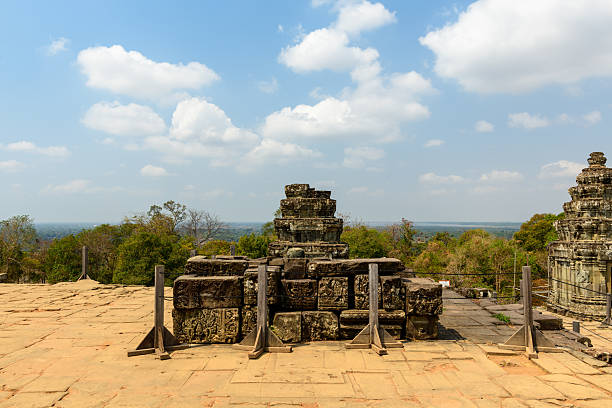 Ancient carved stone blocks form a raised platform on the terrace of Phnom Bakheng temple near Siem Reap, Cambodia. The scene features intricate bas-reliefs, warm daylight, and a backdrop of green forest under a sky dotted with white clouds.