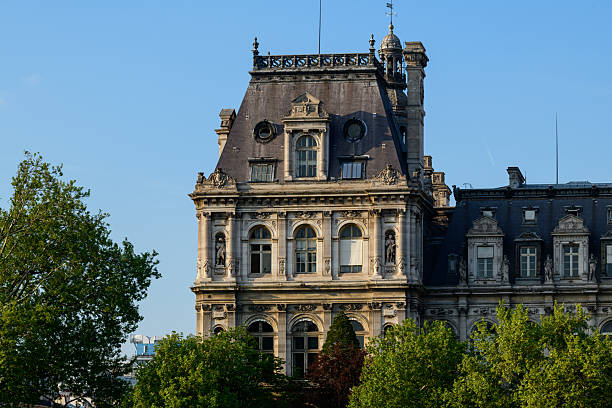 Upper section of the Hotel de Ville in Paris with ornate stonework and arched windows, rising above lush green trees in clear afternoon light.