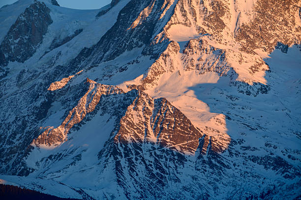 Warm sunrise light casts dramatic orange and pink hues across the snow-covered, jagged slopes of the Mont Blanc massif, highlighting intricate rock textures and deep winter shadows.