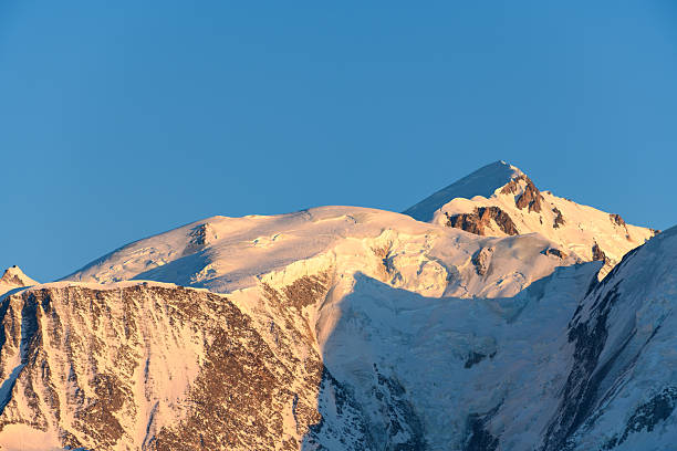 Snow-covered Mont Blanc summit glows with gentle golden hues at sunrise, revealing crisp glacier textures and rugged alpine contours beneath a clear blue sky.