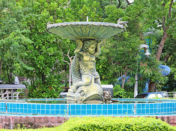 A classical fountain sculpture featuring a cherub holding a large seashell basin, standing on a fish, set in a lush green garden.