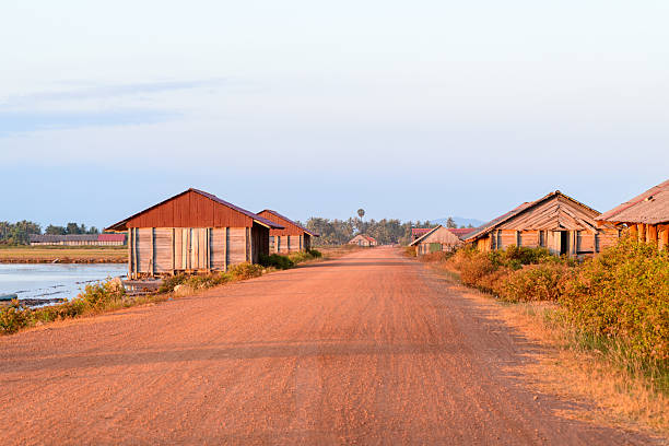 Warm sunlight illuminates rustic wooden salt storage sheds with red metal roofs along a wide red dirt road near Kampot, Cambodia. The tranquil rural scene features open salt ponds, scattered vegetation, and distant palm trees under a pale sky.